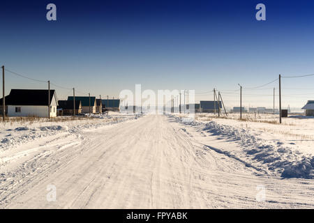 Neve invernale coperto strada conduce attraverso un villaggio domestico verso l'orizzonte. Crystal clear blue sky pende overhead. Il usurato r Foto Stock