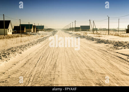 Neve invernale coperto strada conduce attraverso un villaggio domestico verso l'orizzonte. Crystal clear blue sky pende overhead. Il usurato r Foto Stock