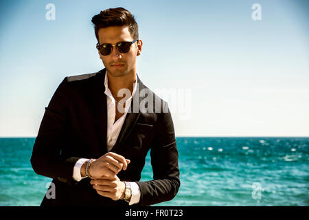 Giovane uomo bello in abito classico sulla spiaggia tenendo gli occhiali da sole mentre guardando lontano. Le onde del mare sullo sfondo Foto Stock