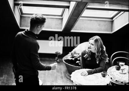 Giovane bella coppia elegante in un abito rosso nella storia di amore al vintage cafe con grandi finestre a tetto, ragazzo dà un fiore Foto Stock