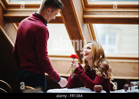 Giovane bella coppia elegante in un abito rosso nella storia di amore al vintage cafe con grandi finestre a tetto, ragazzo dà un fiore Foto Stock