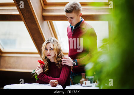 Giovane bella coppia elegante in un abito rosso nella storia di amore al vintage cafe con grandi finestre a tetto, ragazzo dà un fiore Foto Stock