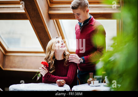 Giovane bella coppia elegante in un abito rosso nella storia di amore al vintage cafe con grandi finestre a tetto, ragazzo dà un fiore Foto Stock