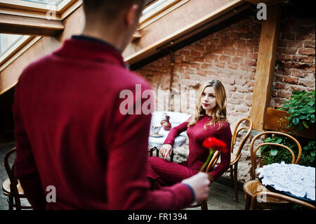 Giovane bella coppia elegante in un abito rosso nella storia di amore al vintage cafe con grandi finestre a tetto, ragazzo dà un fiore Foto Stock