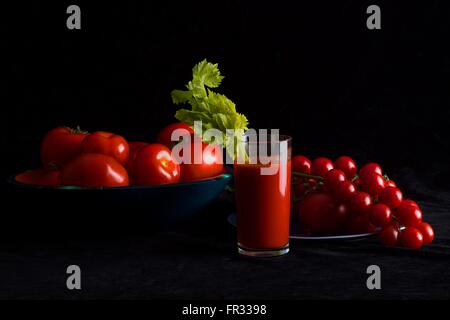 Bicchiere di succo di pomodoro con un gambo di sedano accanto a bocce di pomodori freschi di varie dimensioni su uno sfondo nero. Foto Stock