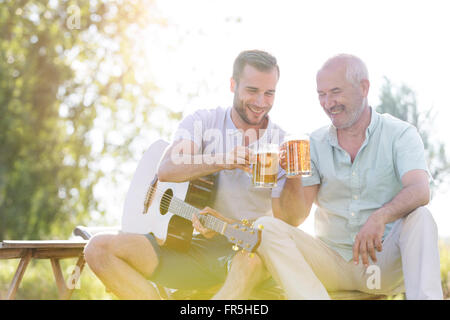 Padre e figlio adulto tostare boccali da birra e suonare la chitarra all'aperto Foto Stock