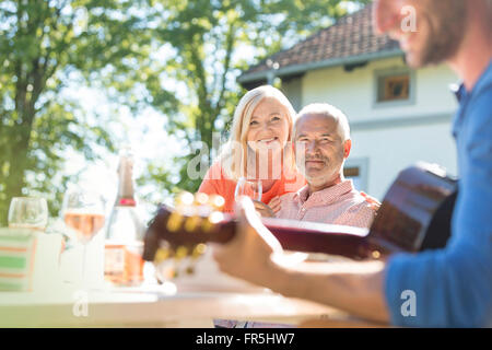 Coppia senior di bere vino e guardare il figlio adulto a suonare la chitarra sul patio soleggiato Foto Stock