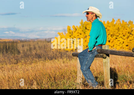 Maschio con cappello Cowboy Straw e occhiali da sole seduti in cima A Una fence di legno guardando in lontananza con alberi colorati in autunno nel Backgrou... Foto Stock
