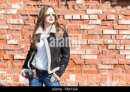 Bella ragazza europea in occhiali da sole con fotocamera rosso contro un muro di mattoni. Felice giovane donna alla giornata di sole Foto Stock