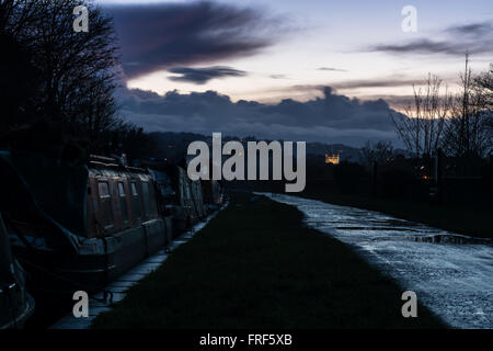 Tramonto su Kennet and Avon Canal. Gli uomini siedono sui loro stretto in barca con un cane e un Viandante è visibile come un fantasma sul percorso di traino Foto Stock