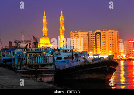 Giù nel porto di Sharjah con Dhows davanti e moschea illuminata sul retro, Dubai, UAE Foto Stock