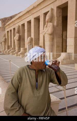 Una guida locale sorsi acqua vicino i colossi dei faraoni presso gli antichi egiziani Tempio di Hatshepsut vicino alla Valle dei Re, Luxor, la Valle del Nilo in Egitto. Il tempio mortuario della Regina Hatshepsut, Djeser-Djeseru, si trova sotto le scogliere a Deir el-Bahari ("il monastero del Nord'). Il tempio mortuario è dedicata al dio sole Amon-Ra ed è considerato uno dei monumenti di incomparabile di antico Egitto.' Il tempio era il luogo del massacro di 62 persone, la maggior parte dei turisti, da islamici il 17 novembre 1997. Foto Stock