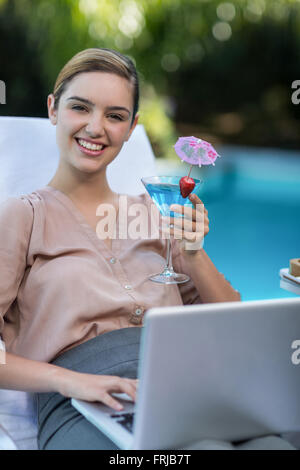 Imprenditrice tenendo un bicchiere di martini durante l'utilizzo di laptop Foto Stock