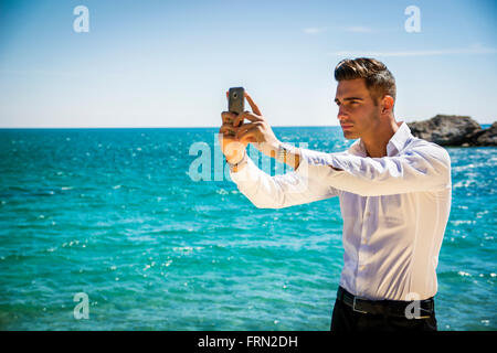 Vista laterale di un elegante giovane uomo prendendo Selfie foto in spiaggia in piedi sotto il calore del sole. Foto Stock