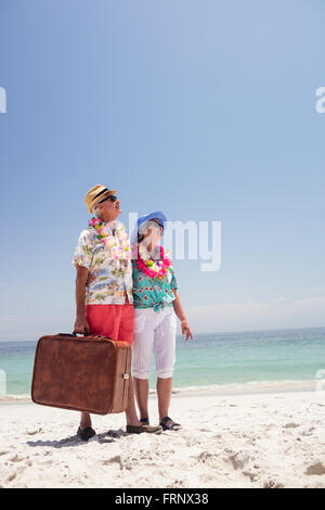 Felice coppia senior permanente sulla spiaggia con la valigia Foto Stock