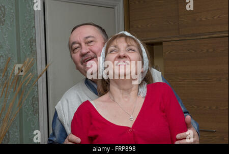 Uomo più anziano in bianco Gilet di maglia è abbracciando il suo sorridente ragazza in rosso brillante camicetta con una scollatura mettendo le mani per la sua Foto Stock