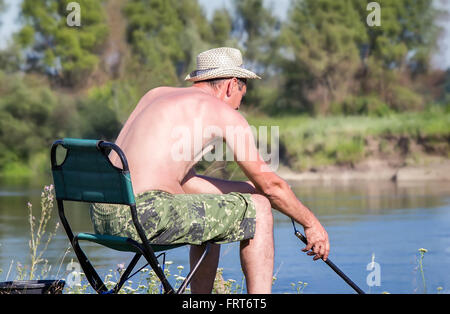 Sulle rive del fiume su una sedia pieghevole si siede un pescatore con canna da pesca e la cattura del pesce. Foto Stock