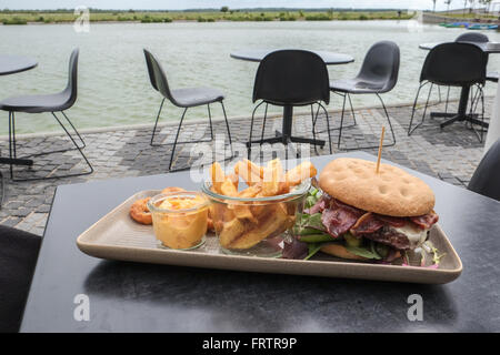 Hamburger di manzo su una piastra quadrata su una tavola nera, con patatine fritte, dip e anelli di cipolla, servita in un caffè all'aperto dall'acqua Foto Stock