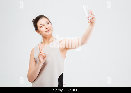 Felice piuttosto giovane donna business tenendo selfie con lo smartphone e mostra il gesto di pace su sfondo bianco Foto Stock