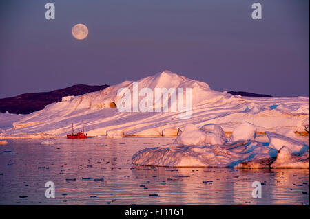 Luna piena al di sopra di barca da pesca nella parte anteriore di iceberg nel crepuscolo, Ilulissat icebergs, Ilulissat, Qaasuitsup, Groenlandia Foto Stock