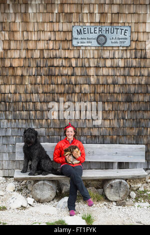 Escursionista femmina seduto con due cani su un banco di lavoro prima del segno della Meilerhütte baita di montagna alla gamma di Wetterstein, Bavaria Foto Stock