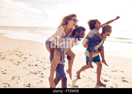 Due giovani uomini dando loro fidanzate piggyback passeggiate sulla spiaggia. Allegro giovani amici godendo di estate sulla spiaggia. Foto Stock