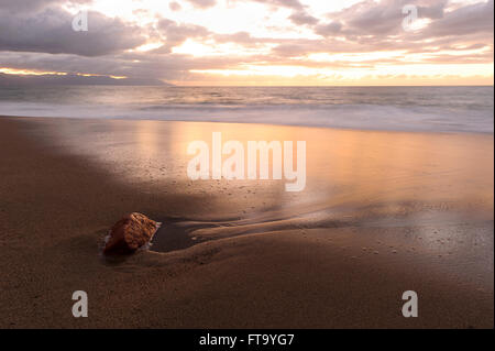 Tramonto sulla spiaggia è un'immagine dettagliata di un morbido tramonto di sabbia con una nube riempiva cielo e una solitaria spiaggia rock in riva al mare. Foto Stock