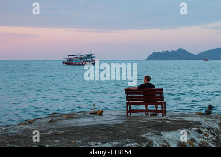 Lonely Man si siede su un banco di lavoro sulla costa guardando il mare. Foto Stock