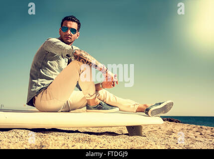 Elegante bel giovane uomo specchiato in occhiali da sole seduto sulla tavola da surf in spiaggia in presenza di luce solare Foto Stock