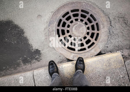 Urbanite man in black nuovi fulgidi scarpe di cuoio in piedi sul ciglio della strada vicino a rusty tombino fognario Foto Stock
