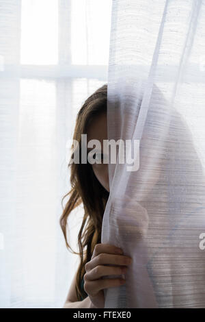 Grave giovane donna di nascondersi dietro le tende da finestra Foto Stock