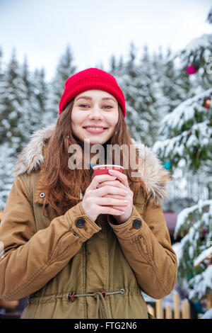 Ritratto di Allegro attraente giovane donna di bere il caffè caldo nei pressi di albero di Natale decorato nella foresta di inverno Foto Stock