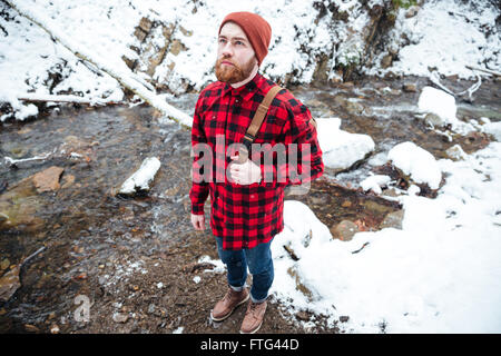 Vista superiore del pensieroso barbuto Giovane Uomo in camicia a scacchi e hat in piedi vicino al fiume di montagna in inverno Foto Stock