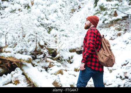 Grave barbuto giovane uomo in plaid shirt e jeans con zaino a piedi nella foresta di inverno Foto Stock