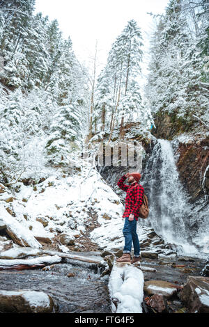 Attraente barbuto Giovane Uomo in camicia a scacchi e jeans camminare vicino a cascata in montagna in inverno Foto Stock