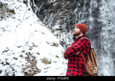 Riflessivo barbuto Giovane Uomo in camicia a scacchi e hat in piedi con le braccia incrociate vicino alla cascata di montagna in inverno Foto Stock
