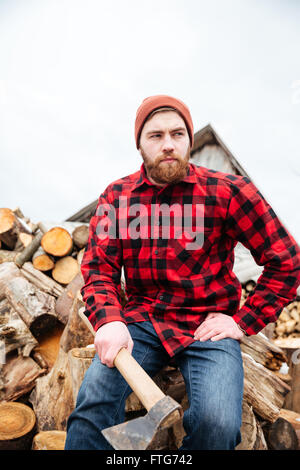 Grave giovane uomo bello in camicia a scacchi seduta su log e azienda ax Foto Stock