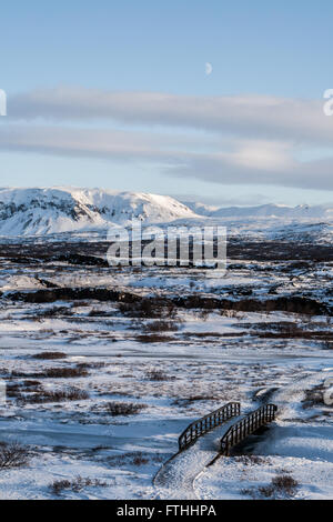 Coperta di neve ponte attraverso il fiume a Thingvellir National Park in inverno con montagne innevate e la luna nel cielo blu chiaro Foto Stock