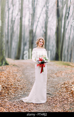 Felice bella bionda sposa sulla foresta di autunno con bouquet di fiori di colore rosso Foto Stock