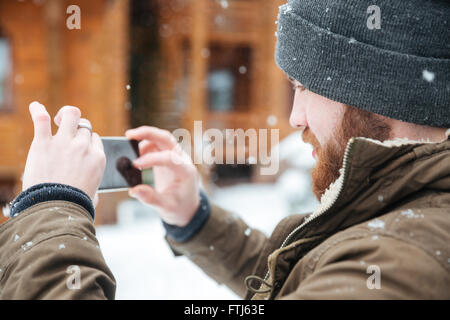 Concentrato uomo barbuto a scattare foto con il cellulare in caso di neve in inverno Foto Stock