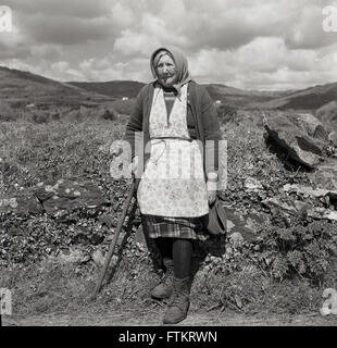1950s, foto storica di J Allan Cash di una donna irlandese matura che indossa un grembiule o una pinafore sopra una gonna di lana, un cardigan e una sciarpa sopra la testa, in piedi con un bastone, da un hedgerow, Irlanda rurale. Foto Stock