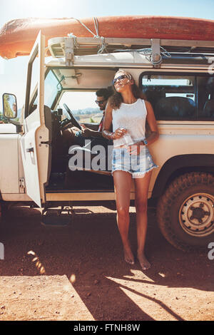 Coppia giovane sulla vettura roadtrip. L uomo e la donna prendendo una pausa dal viaggio. Donna in piedi al di fuori della vettura con una bottiglia di w Foto Stock