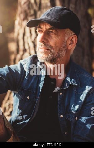 Close up ritratto di uomo maturo che indossa il cappuccio che guarda lontano mentre è seduto da un albero. L'uomo anziano con la barba all'aperto su un giorno di estate Foto Stock