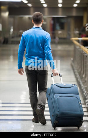 Giovane uomo tirando la valigia in aeroporto moderno terminale. Viaggiare ragazzo indossa smart casual style vestiti a piedi Foto Stock