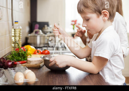 Sorridente piccolo ragazzo sbattere le uova nella ciotola sul tavolo. Cucina di famiglia dello sfondo. Foto Stock