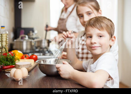 Sorridente piccolo ragazzo con mamma e mia sorella sbattere le uova nella ciotola sul tavolo. Cucina di famiglia dello sfondo. Foto Stock