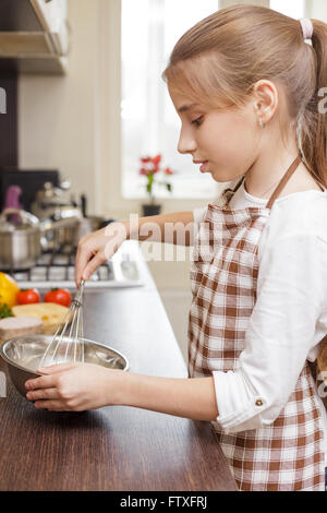 Piccola ragazza adolescente in grembiule sbattere le uova nella vaschetta bianca in cucina Foto Stock