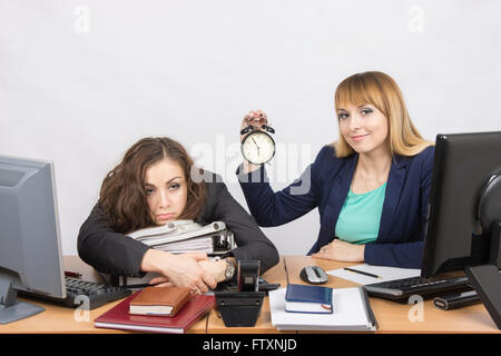 Due ragazze in ufficio alla fine della giornata, uno con un sorriso, tenendo un orologio, un altro affaticati giace sulle cartelle Foto Stock