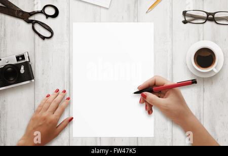 Donna di scrivere con la penna rossa su isolato, libro bianco. Caffè, bicchieri, fotocamera, notepad matita su un tavolo di legno. Vista dall'alto. Foto Stock