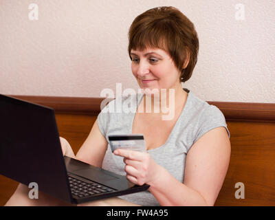 Ritratto di attraente donna matura seduta alla camera da letto con il suo computer portatile e la holding carta bancaria in mano Foto Stock